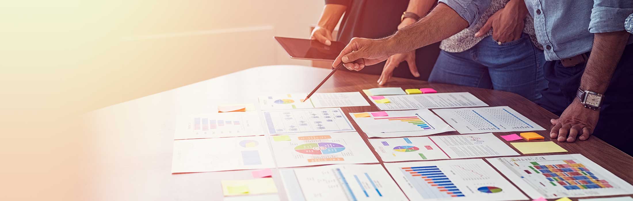 Close-up of a hand holding a pencil and pointing to graphs on a table while analysts brainstorm and discuss reports.